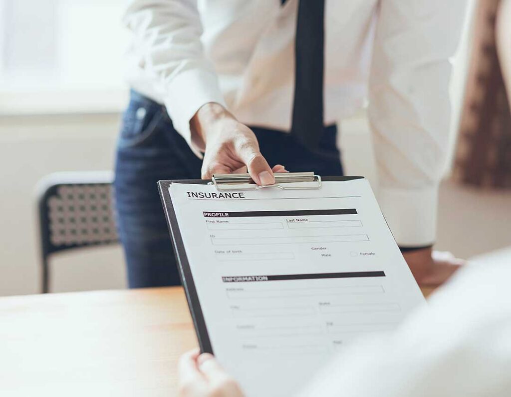 A person reviews insurance documents at a kitchen table with a family and a laptop nearby.